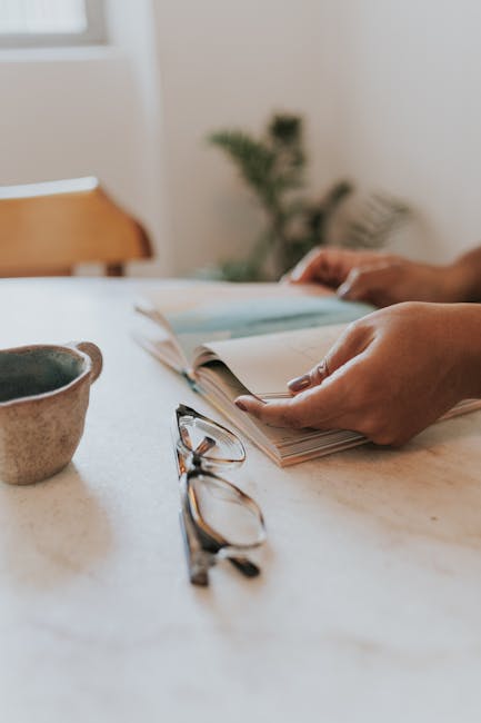 A serene morning scene with hands holding a book, a coffee mug, and glasses on a marble table.